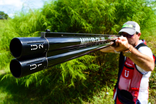Close-up of a shooter holding an over-and-under shotgun at a gun club, focusing on the extended clay target chokes at the end of the barrels.